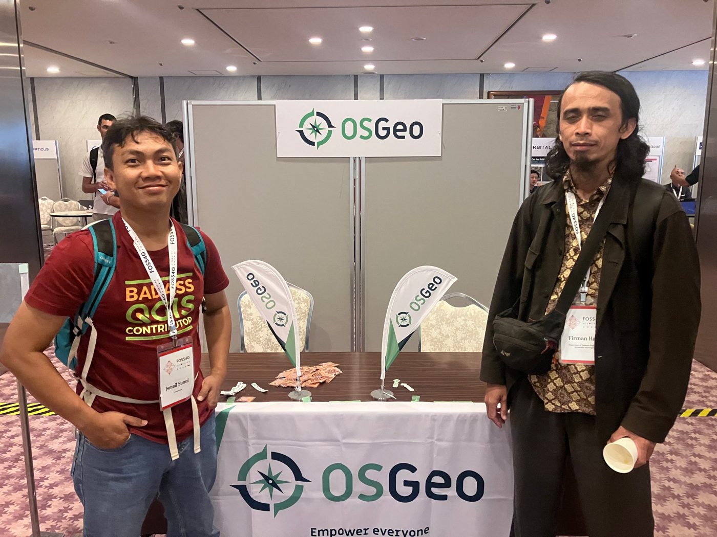 Two participants standing beside the OSGeo booth at FOSS4G 2026